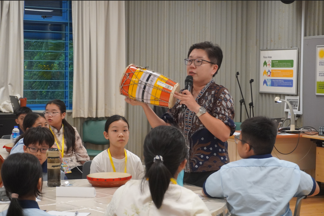 An instructor holding up a colorful traditional drum while speaking to seated students.